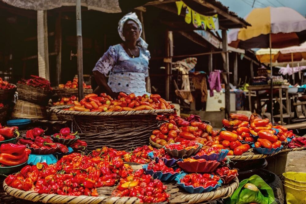 A market vendor stands behind baskets of fresh chili peppers and tomatoes